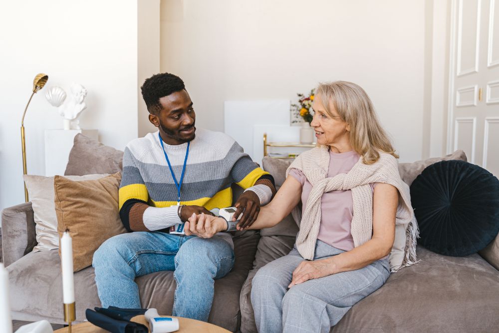 A healthcare worker sits beside a person on a sofa and measures blood pressure using an arm cuff and monitor in a home living room setting. A blood pressure monitor is also visible on a coffee table in the foreground.