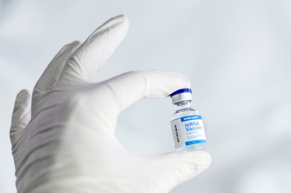 A white-gloved hand holds a small vial labelled “mRNA Vaccine COVID-19”, against a softly blurred pale background.
