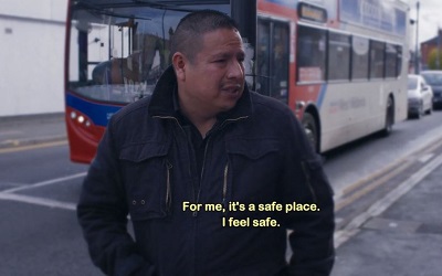 This photo shows a screen shot from the film - Jose, the main individual featured in the film, is walking along a street with a large red bus about to drive past
