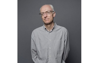 Photo shows Dr Rob Macmillan in a grey shirt on a grey background. He is looking away from the camera at something. He has short white hair and glasses