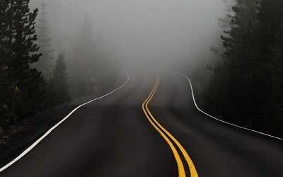 Photo of misty road with a bend up ahead - pine trees sit either side. The image creates a sense of the unknown, uncertainty but also exciting adventure. By Katie Moum on Unsplash