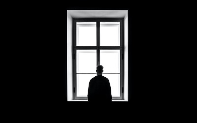 sasha-freemind-Pv5WeEyxMWU-unsplash Photo shows a man standing in a window in a dark room. He looks out. The photo creates a sense of sadness and loss. The rest of the room is black.