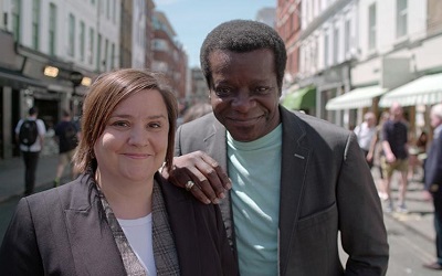 Stephen K Amos and Susan Calma, the presenters, stand next to each other smiling in a street