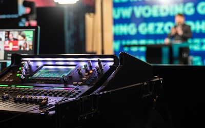 A mixing desk sits next to a screen infront of a stage where a broadcast is taking place