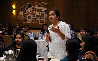A female stands in a room of guests wearing a white shirt. She holds a microphone and is asking a question to the speaker who is out of shot 