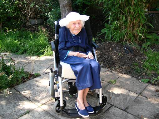 Patricia Collen sitting in a wheelchair, on a sunlight dappled patio, smiling cheekily at the camera