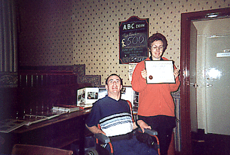 A colour photo of a woman standing with a certificate in her hands, and a man in a wheelchair beside her