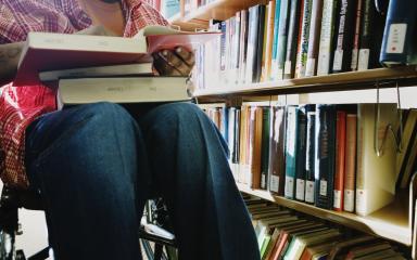 A student in a wheelchair has a pile of books resting in their lap. A stack of books can be seen behind them.