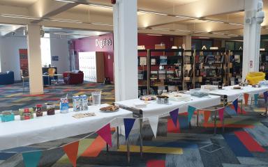 A series of tables are set out on the ground floor of the library building, with a variety of cakes and baked goods placed on them.