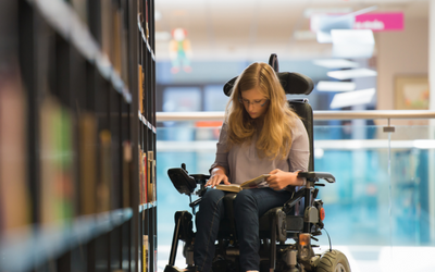 A woman in a wheel chair reading a book near a bookshelf in a library