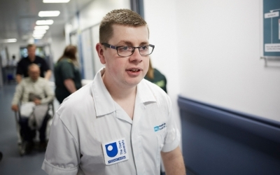 A male nurse in a hospital corridor, wearing a white nurse tunic and glasses