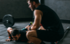 A man in a gym, dressed in black, watching over someone doing push ups