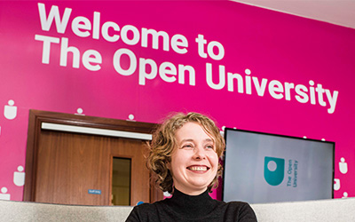 Woman smiling to camera in the OU office reception with 'Welcome to The Open University' in the background