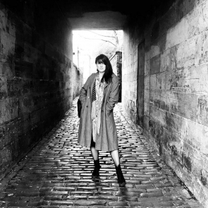 A white woman with long dark hair stands on a cobbled lane. The stone buildings around her form an archway overhead. She is wearing a long coat and boots.