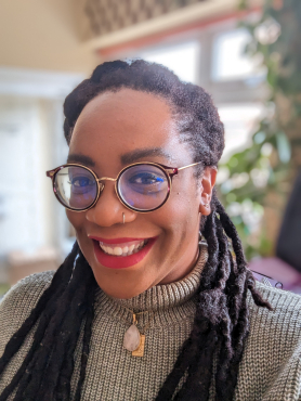 A photo of Black woman smiling, wearing glasses, gold jewellery, and a light green jumper 