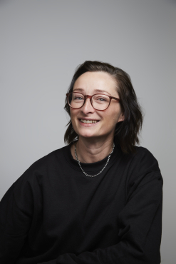 Tracie is wearing a black sweatshirt and silver chain with medium length brown hair and glasses