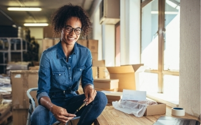 Woman with her own small business, sitting at a desk with a laptop and packaging materials. 