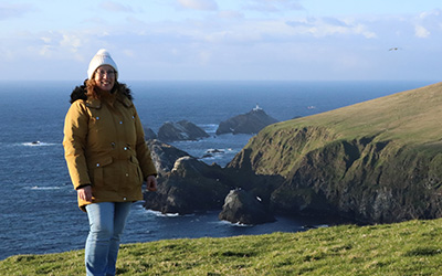 OU graduate Michelle on a Shetland cliffside with the sea, rocks and a lighthouse in the background