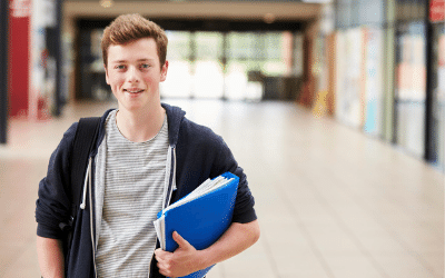 A smiling male student standing in a college building, and holding a file under his arm.