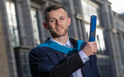 OU graduate Calum Waters pictured in a graduation gown and holding a degree scroll.