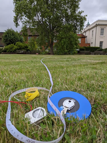 Tape measure and compass lie on the ground with a large tree and buildings in the distance
