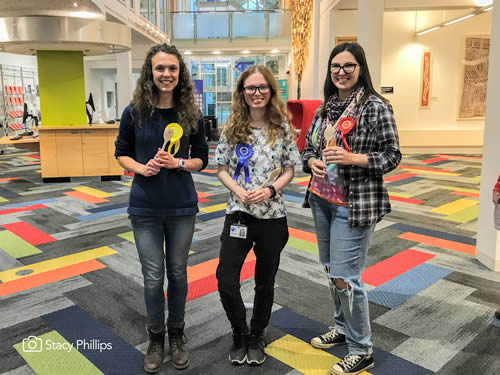 Three female PhD students stand wearing rosettes.