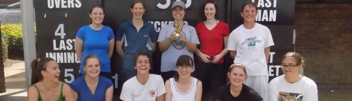 A team of female cricketers stand in front of teh scoreboard holding their trophy.