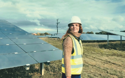 Female Open University student next to solar panels wearing hard hat and hi vis vest
